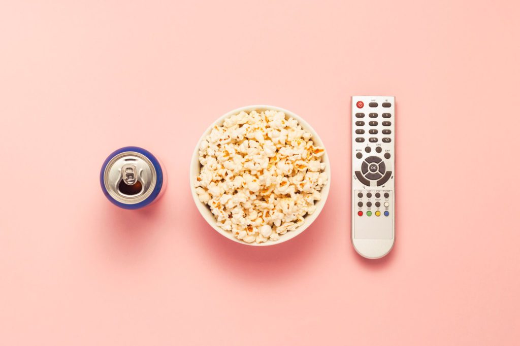 Can of soda, popcorn, and remote getting ready to watch a movie from the list of must-watch Netflix things this summer.