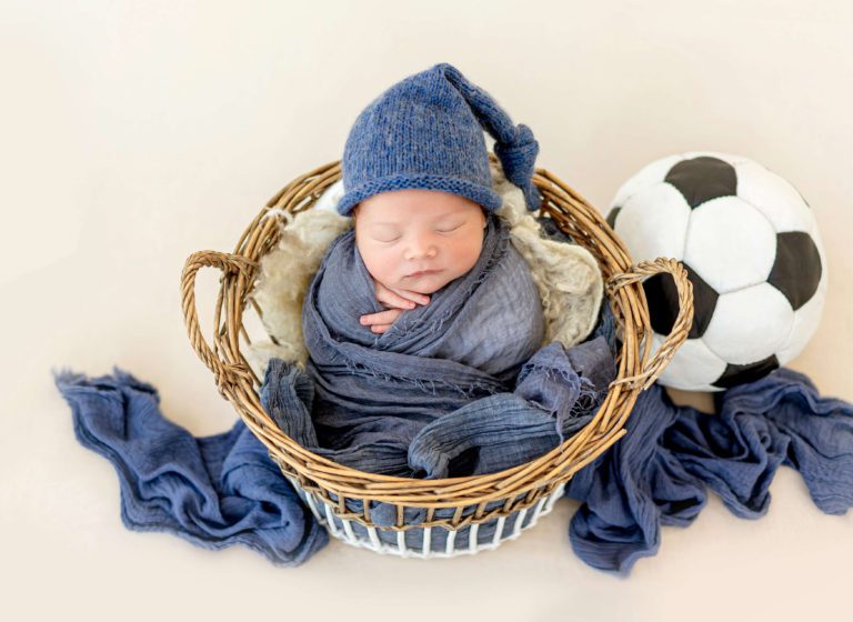 Newborn baby in a basket with a hat on and a soccer ball beside him