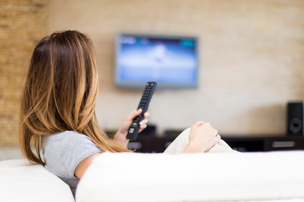 Woman on the couch with a remote in her hand deciding what to watch from her list of must-watch Netflix things this summer.