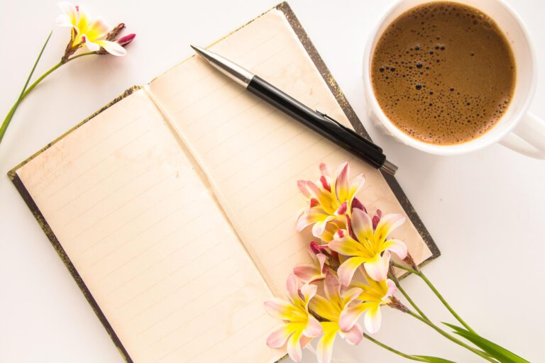 Cup of coffee next to a coffee journal with a pen and flowers