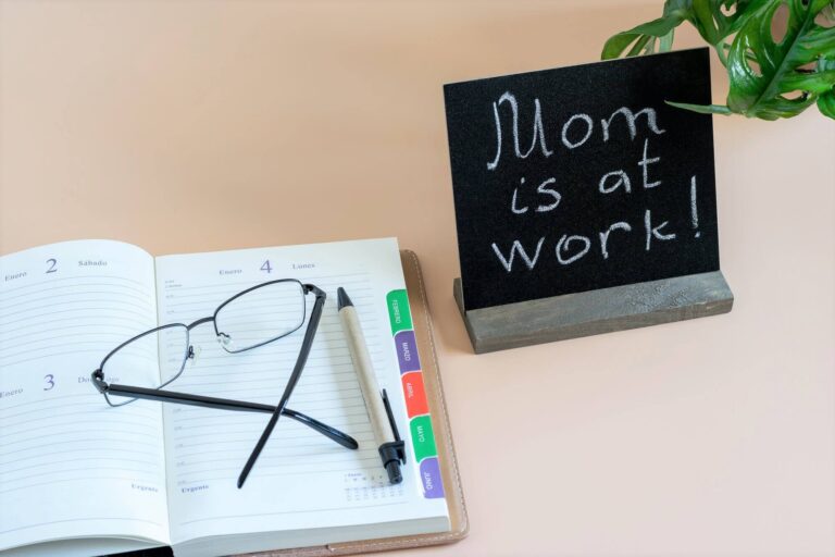 Sign that reads "Mom is at work!" next to a planner with eyeglasses on top.