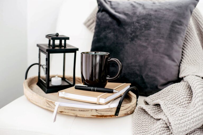 A cup of coffee on top of a journal filled with notes about different coffee roasts. The journal is on a wood tray next to a lantern with a pillow and blanket in the background.