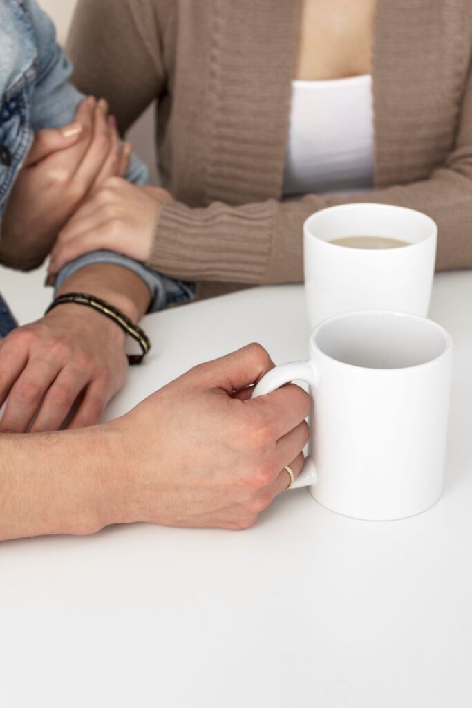 Hands and arms of two coffee moms sitting and drinking out of white coffee mugs.