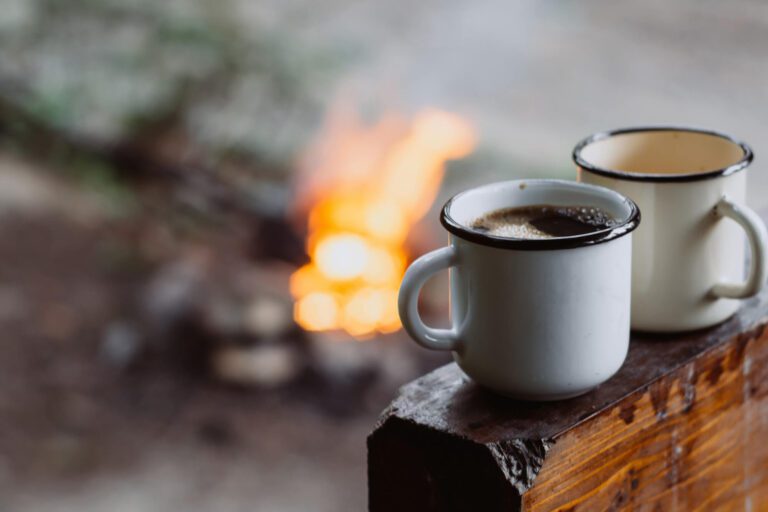 Two mugs of coffee sitting on a log next to a campfire in the background.