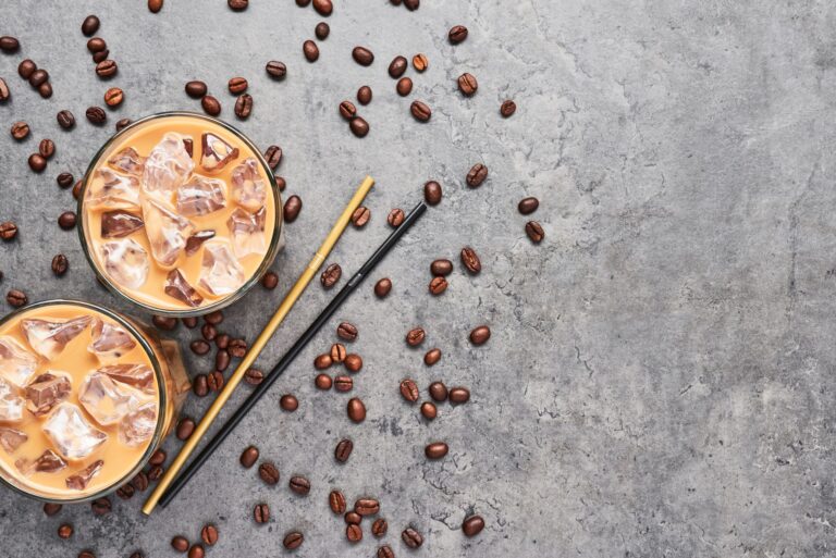 Iced coffee glasses in a flatlay display on a grey background with coffee beans scattered around and two straws.