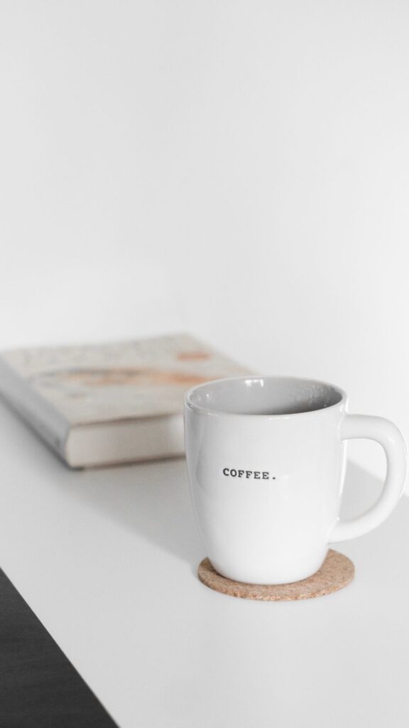 A cup of coffee on a coaster on top of a white desk with a book in the background.