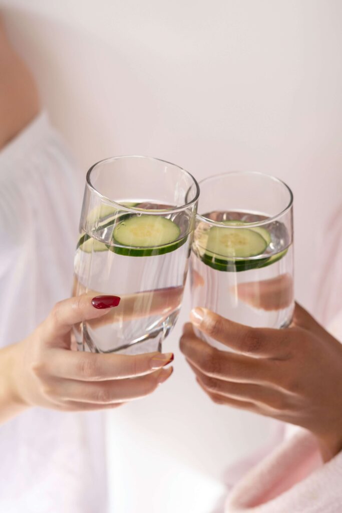 Two women holding glasses of water with cucumber slices floating on top.