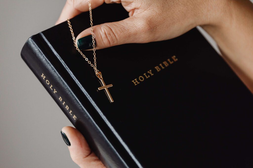 A black Bible with gold lettering in the hands of a woman with a gold cross necklace on top of the Bible.