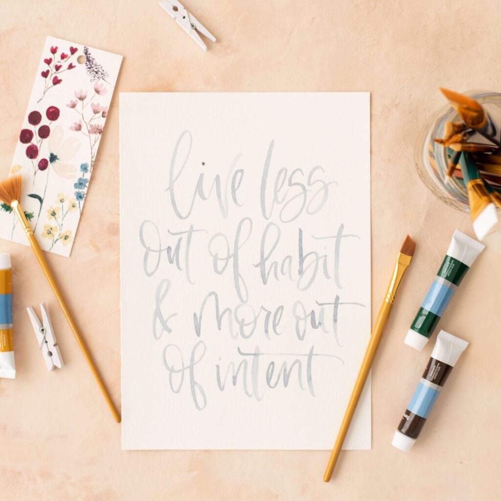 A watercolor of a quote that says, "live less out of habit and more out of intent" with art supplies on the sides.