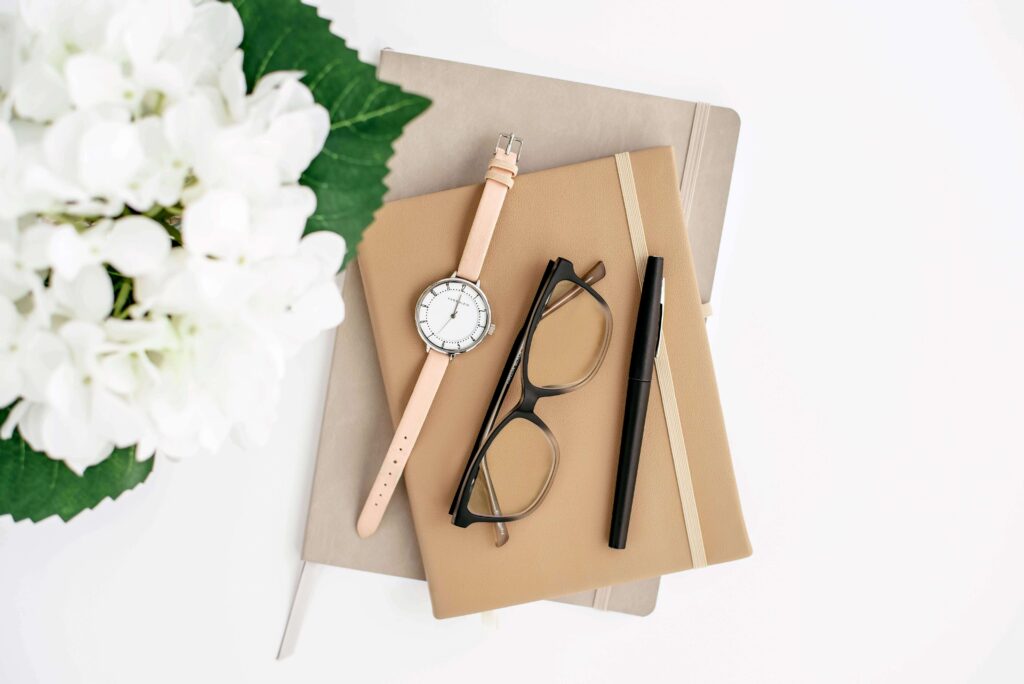 Flatlay image of journals with a watch, glasses, and pen on top next to white flowers.