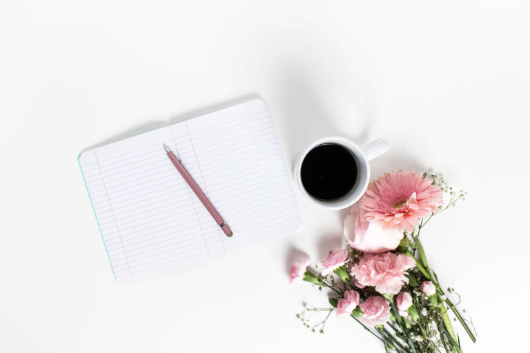 A journal with a pen on top next to a cup of coffee and flowers