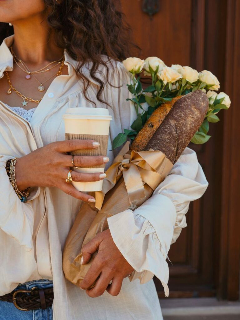 A woman holding bread, flowers, and a cup of coffee in a to-go mug.